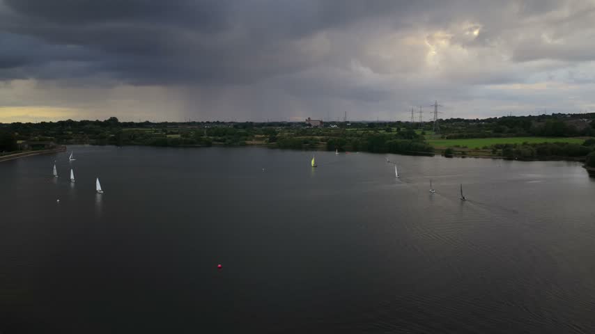 Aerial video  showing a fleet of sailing dinghies gliding across the dark waters of Elton Reservoir alongside the curved stone dam embankment under stormy skies in Bury.
