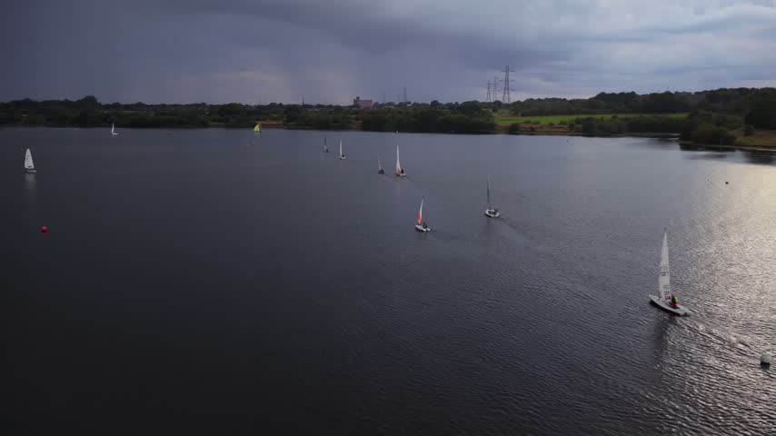 Aerial video  showing a fleet of sailing dinghies gliding across the dark waters of Elton Reservoir alongside the curved stone dam embankment under stormy skies in Bury.