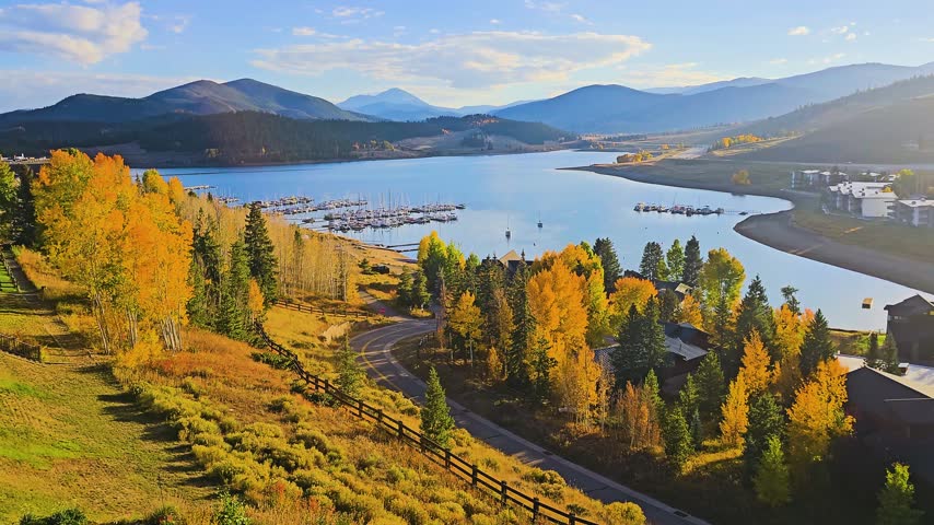 Panning view over a mountain lake marina in autumn, with docked boats, calm blue water, golden aspen trees, winding road, forested hills, and distant peaks under clear sky in sunlight.