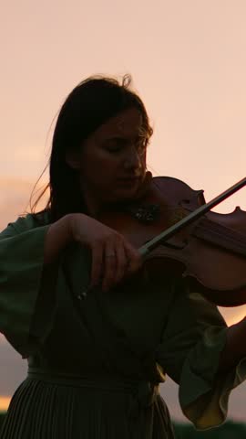 Graceful violinist performing at sunset, silhouetted against a glowing sky while playing soulful music outdoors in a peaceful, contemplative natural setting