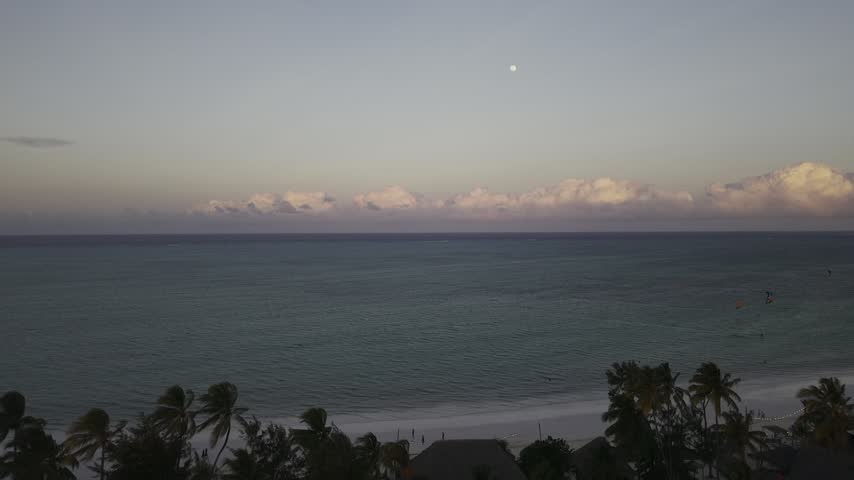 Drone lowers with view of full moon high over clouds on the horizon above the Indian Ocean on a windy afternoon during sunset in Paje, Zanzibar, Tanzania