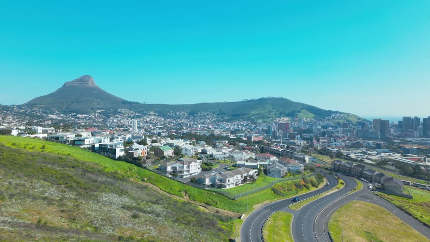 Sweeping drone shot from a grassy hillside overlooking Cape Town’s City Bowl, with Lion’s Head rising on the left, Signal Hill in the middle distance, and the city’s high-rise skyline and harbour.