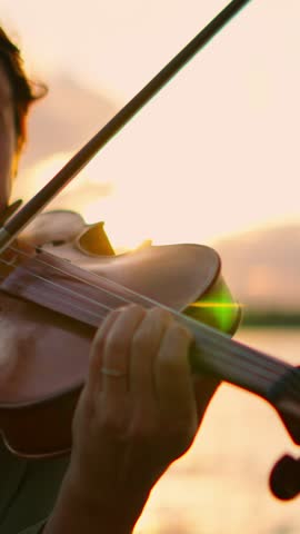 Close-up of violinist playing graceful melody by tranquil waterfront at sunset, warm golden light illuminating strings, fingers, and serene evening atmosphere