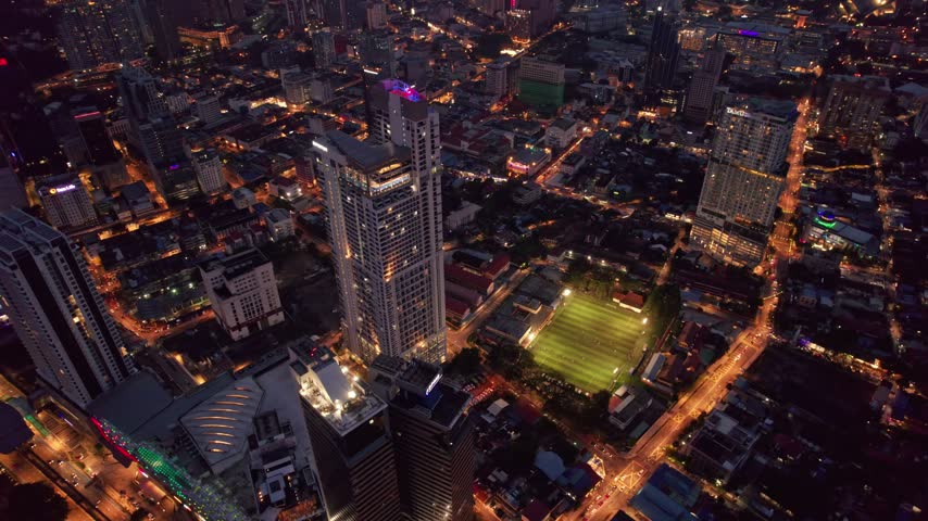 Cinematic nighttime drone footage above Kuala Lumpur showing a brightly illuminated football field surrounded by modern towers, city streets and glowing urban infrastructure.