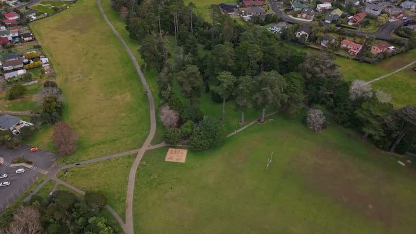 Drone footage showing a large green park, walking paths, mature trees and surrounding residential homes in Henderson, Auckland, New Zealand.