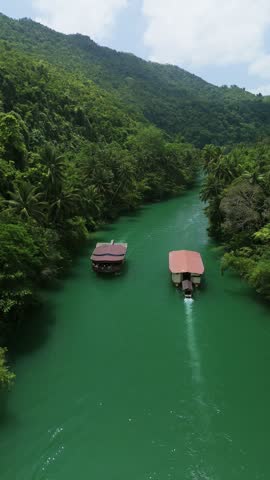 Vertical drone pulls back and ascends over two Loboc River cruise boats traveling upstream through green water and forested hills in Bohol