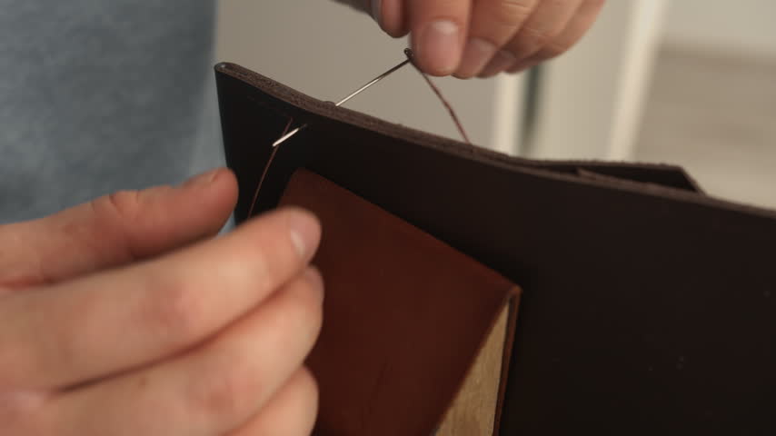 Leather craftsman hands sewing a genuine brown leather wallet with a saddle stitch using a needle and thread in a workshop