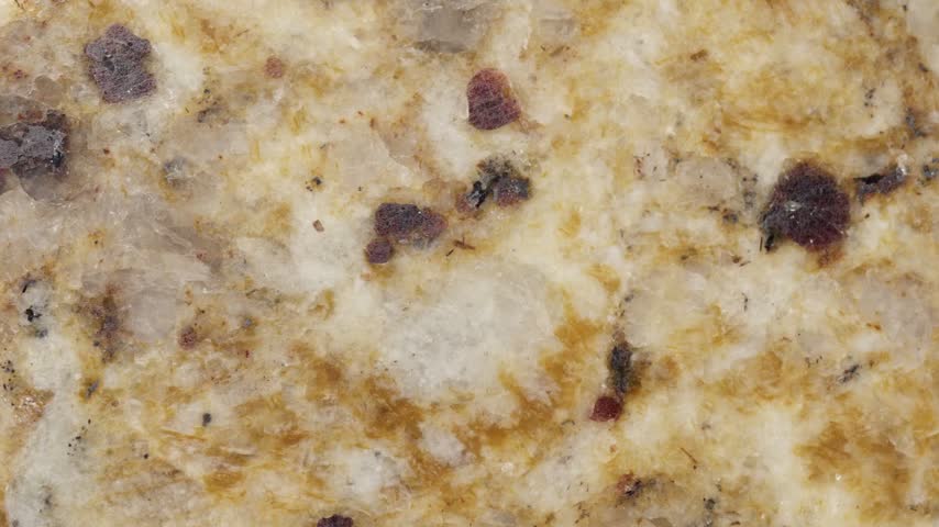 The texture and surface details of the natural granite slowly pan into view. A unique mineral pattern is visible, featuring a blend of cream and brown hues, translucent quartz, and elegant speckles of dark red and black minerals.