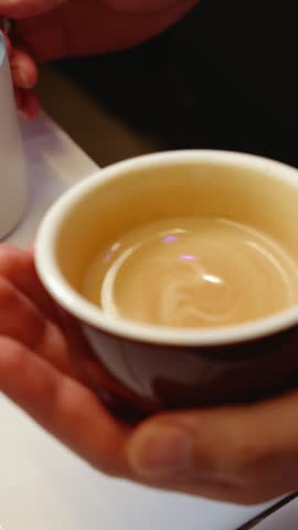 Barista pours steamed milk into coffee to create a latte art pattern. The milk is carefully poured from a pitcher while the cup is held by hand.