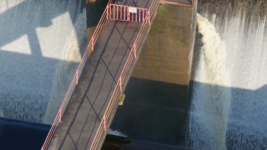 Water flowing over a concrete dam spillway with a pedestrian bridge above