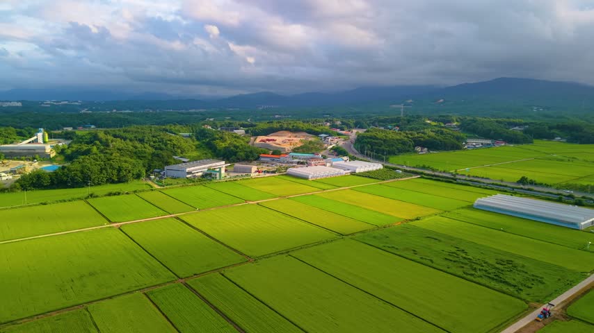 Aerial orbit over rice paddies revealing industrial facility with silo tower nestled among forested hills near Gangneung coastline under cloudy sky