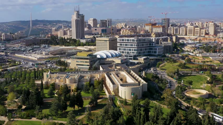 High angle vista of the Supreme Court complex surrounded by green trees with the modern Jerusalem cityscape in the background