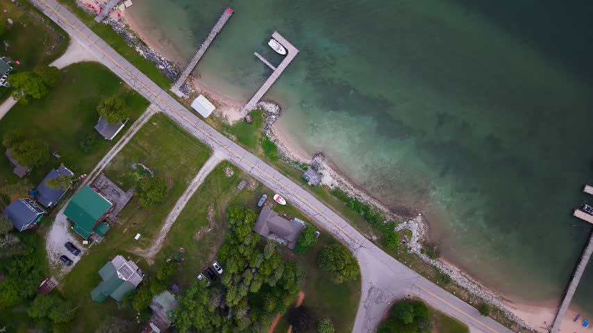 Top down drone view shows a waterfront road homes docks and a small marina along clear Lake Huron water in Michigan.