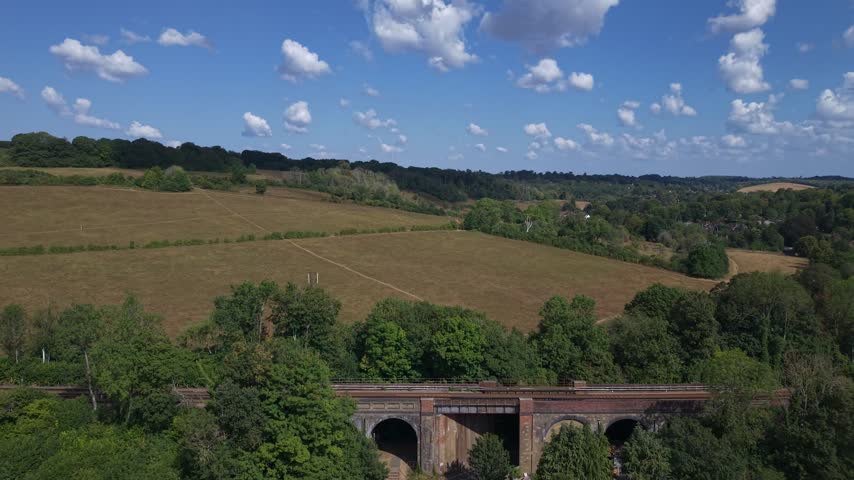 Aerial drone footage flying over the historic Swanage Railway viaduct bridge near Corfe Castle in Dorset, crossing a country road surrounded by green fields and forest under a sunny blue sky.