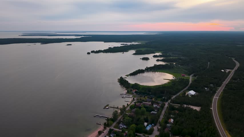 High aerial panorama overlooks a small Lake Huron waterfront community road forest and islands beneath a colorful sunset sky.