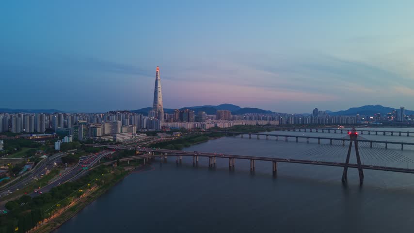 Seoul skyline at blue hour showing Lotte World Tower glowing red beacon and Olympic Bridge lit above Han river as city lights turn on - aerial orbit shot