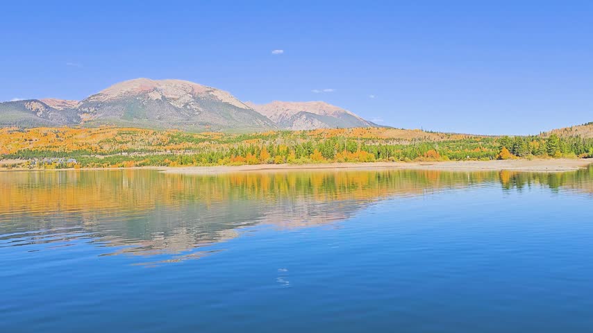 Scenic boat view across a calm alpine lake in autumn, with golden aspen hillsides, evergreen forest, rocky mountain peaks, blue sky, and gentle water reflections drifting past.