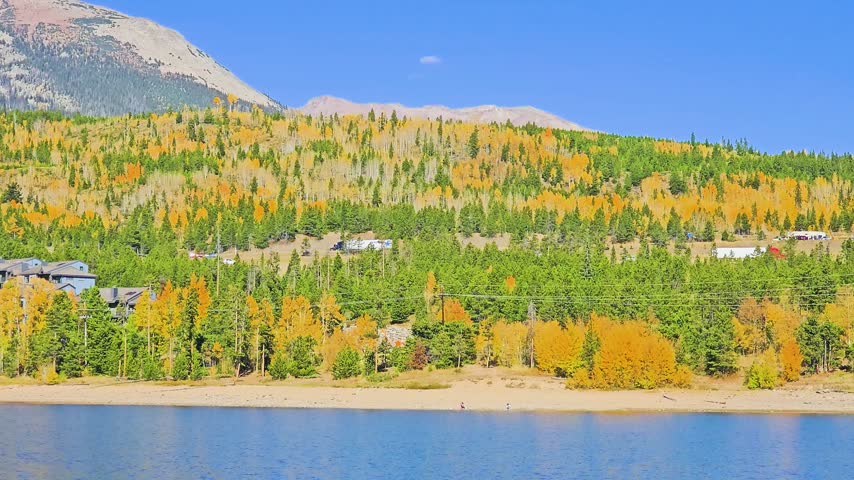 Panning landscape of Dillon Reservoir in fall, with golden aspen forests, rugged Rocky Mountain slopes, lakeside village buildings, reflective water, sunny blue sky, and calm scenery.