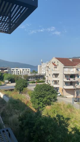 A rightward panning shot showcases apartment buildings, parking lots, and lush green hills under a clear blue sky in Tivat, Montenegro. The panorama sweeps across residential structures with tiled roofs, parked cars, and dense green vegetation, with mountains rising in the background. This daytime view highlights the peaceful Mediterranean-style architecture and natural landscape