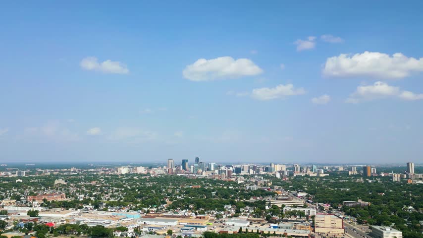Zoomed-in aerial drone shot of downtown Winnipeg with Portage Avenue, office towers, residential neighbourhoods, and city streets beneath a bright summer sky