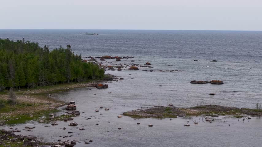 Drone rises over a rocky Lake Huron inlet with scattered reefs and a forested point along the Michigan coast.