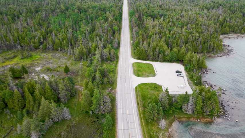 Top down aerial shows a straight rural road and small waterfront turnout cutting through dense Michigan forest beside Lake Huron.