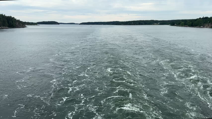 Stern perspective of a large passenger ferry creating a foaming water wake across the Baltic Sea between forested islands in the Stockholm archipelago.