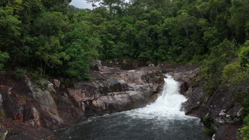 Low-altitude aerial drone footage following a wild mountain stream in Chu Yang Sin National Park, Dak Lak, Vietnam. The camera approaches a rocky section where flowing water drops approximately three meters, forming a striking small waterfall among surrounding cascades. Dense green forest and rugged rocks create a pristine natural setting. The smooth flight highlights the stream