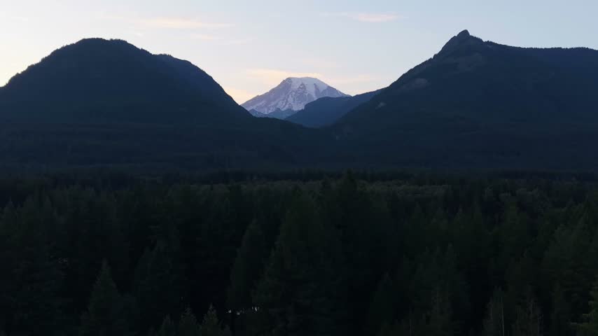 Aerial view of Mount Rainier above a dense forested landscape near Packwood, Washington, with evening light illuminating the surrounding mountain ridges.
