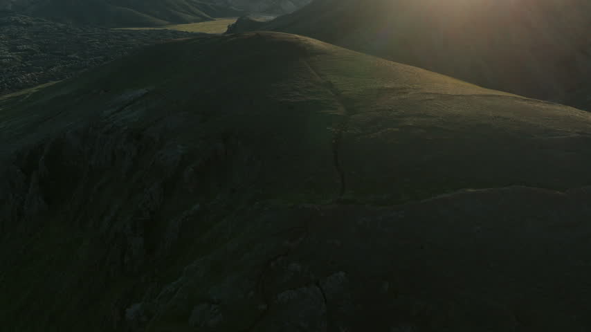 Drone footage of a rolling hillside silhouette glowing at sunset, warm sunlight flaring at the top of the ridge.