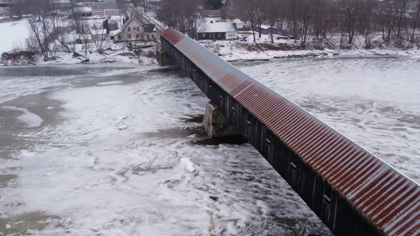 Aerial drone flying alongside the historic Cornish-Windsor Covered Bridge over the frozen Connecticut River in Windsor, Vermont, with the structure offset right and light snow blowing.