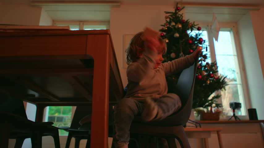 Young child climbs down from a chair next to a wooden dining table, extending one leg toward the floor as Christmas decorations and large windows fill the background