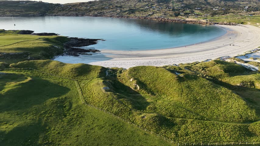 Aerial drone footage overlooking the sheltered waters and sweeping white sandy beach of Dog’s Bay near Roundstone, County Galway, Ireland. Rolling green dunes, rocky coastal terrain, a sailboat and scattered houses surround the turquoise Atlantic bay in warm evening sunlight.