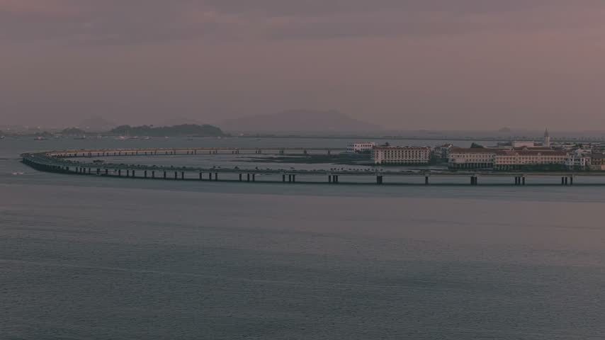 Aerial pan right across a long coastal highway and waterfront in Panama during sunset, surrounded by calm sea and distant urban landscape.
