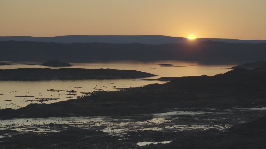 Continuous 30-second real-time landscape clip capturing the Arctic sun slowly dipping below distant tundra hills, turning the sky and water gold over Frobisher Bay.
