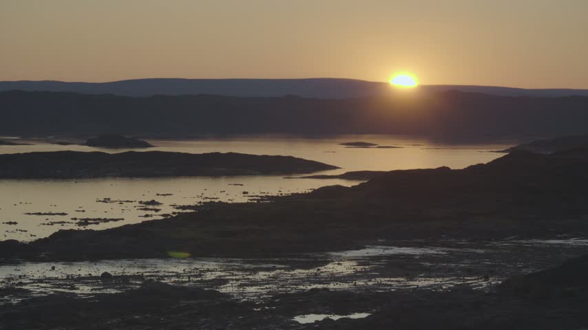 Silhouette landscape capturing the low summer sun hovering over distant tundra hills and casting deep orange reflections across the bay