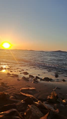 Vertical video of golden sunset over Aegean Sea coast with windy weather. View of bright glowing sun dipping towards horizon over wavy sea waters and pebbled beach, with warm sunlight reflection on wet sand and sea waves. Perfect for nature background, coastal summer sunset, travel, and windy outdoor atmosphere.