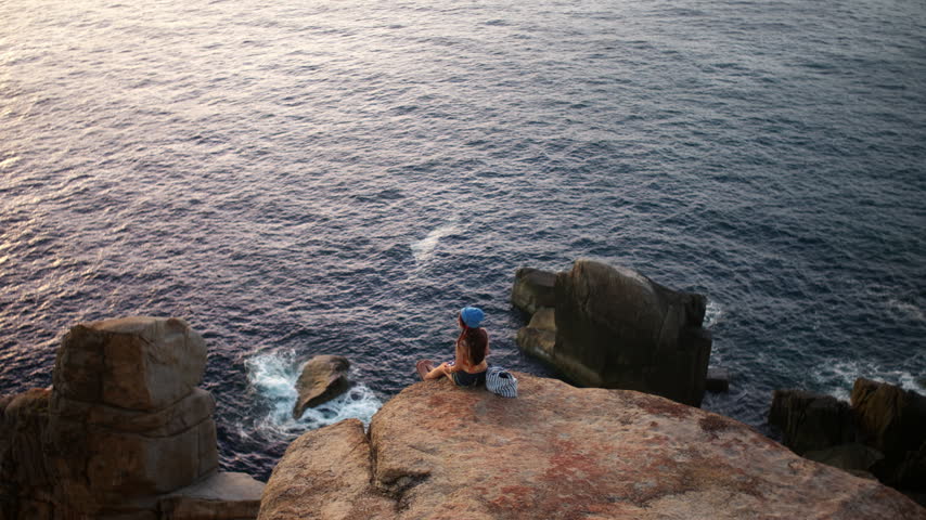 Solo young woman girl sits on a high rocky ledge above ocean water surrounded by rugged coastal rocks, staring out with admiration and longing as she thinks about her future
