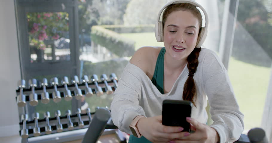 Woman sitting on home bike holding phone, wearing loose top, laughing after funny notification. Female, fitness, elliptical, headphones, smartwatch, dumbbells, window