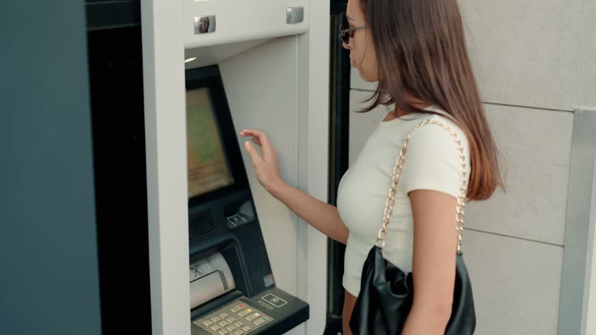 Young brunette woman in sunglasses using an automated teller machine, inserting her credit card to withdraw cash and complete her financial transaction at an outdoor bank terminal