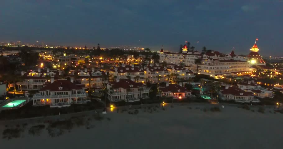 Aerial view of Coronado coastal neighborhoods glowing with city lights at night, with the beach and illuminated waterfront visible nearby.