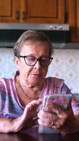 Serious senior woman with short hair and glasses sitting at a table in her kitchen, intently focused on scrolling and tapping on her mobile phone, staying connected with technology