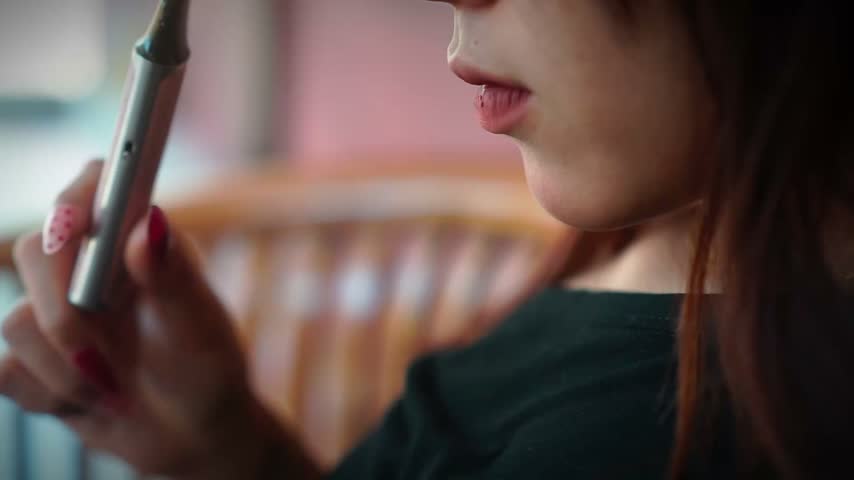 Detail shot of young asian woman holding silver vape device, inhaling and exhaling vapor. Concept of smoking alternative, nicotine addiction, or modern lifestyle habit.