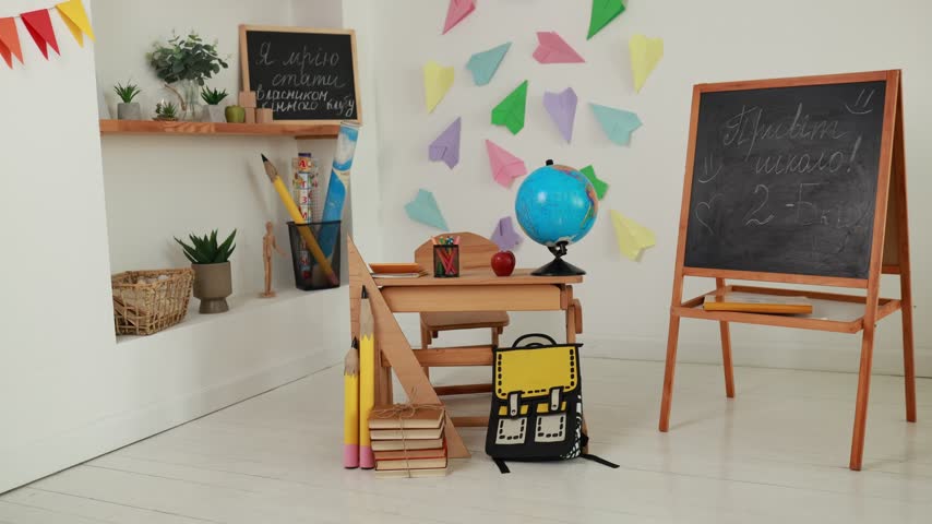 Empty primary interior school classroom setup with wooden desk, globe, apple, chalk board easel. Back to school concept, colorful paper airplanes wall decor, study background, and school supplies.