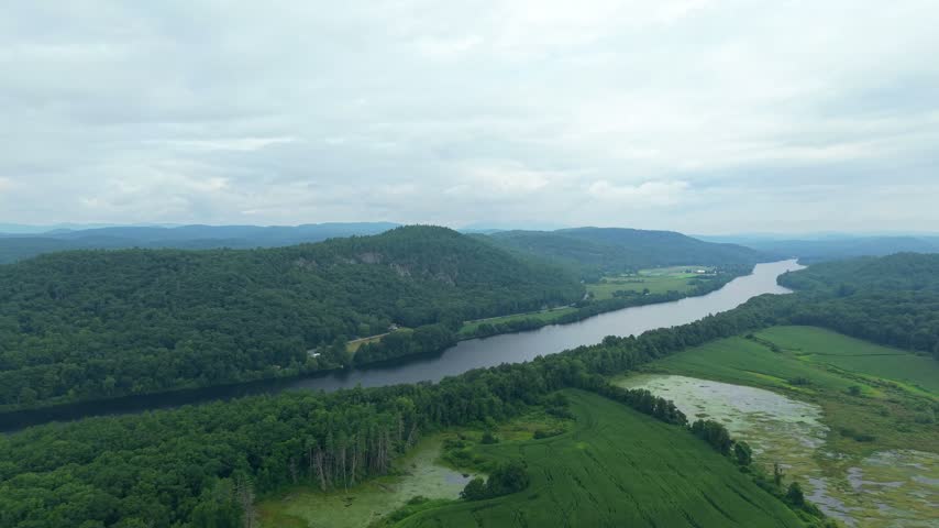 Aerial drone video over the Connecticut River in Charlestown, New Hampshire, flowing through lush green farm fields, wetlands, and forest-covered hills under a soft overcast sky.