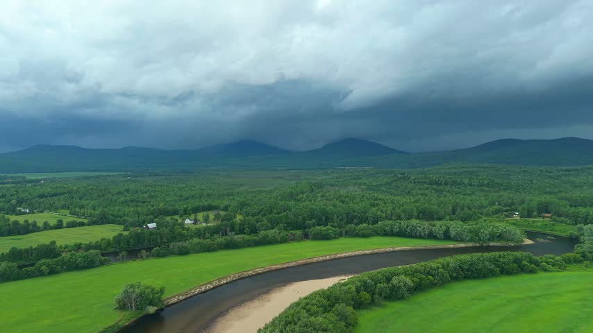 Groveton, New Hampshire, high aerial drone video over lush green forests, farmland, and a winding river bend, set against distant mountains under a dark, dramatic stormy sky.