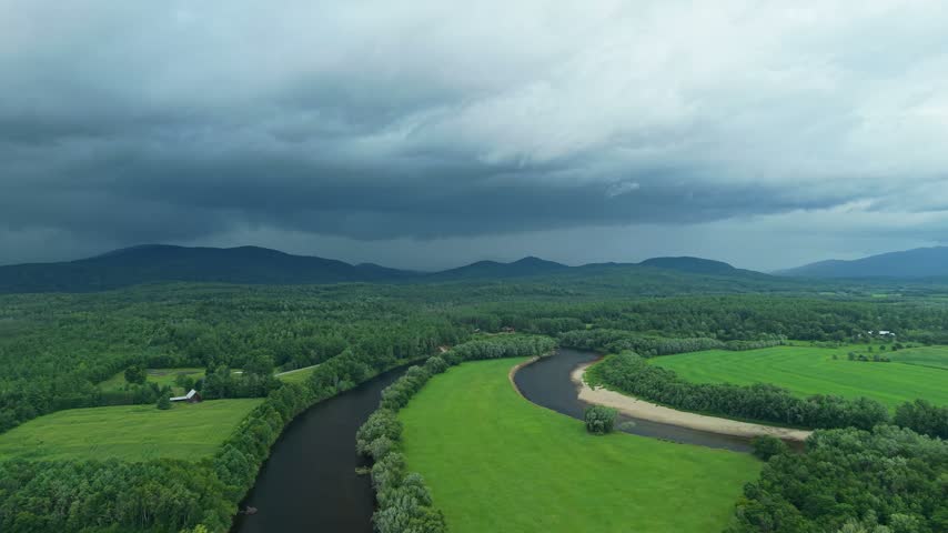 Groveton, New Hampshire, high aerial drone video over lush green forests, farmland, and a winding river bend, set against distant mountains under a dark, dramatic stormy sky.