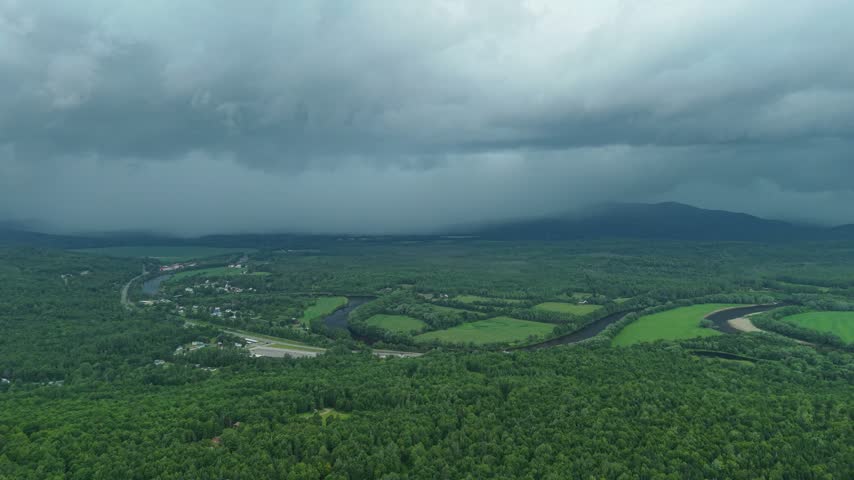 Groveton, New Hampshire, high aerial drone video over lush green forests, farmland, and a winding river bend, set against distant mountains under a dark, dramatic stormy sky.