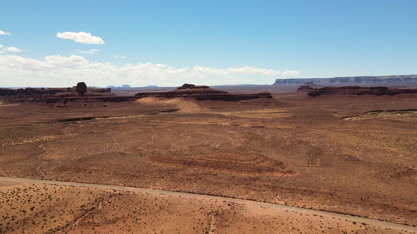 Aerial drone view of Valley of the Gods, a remote sandstone basin near Mexican Hat in San Juan County, southeastern Utah, USA. This aerial drone video reveals a vast desert landscape scattered with towering red buttes, isolated mesas and dramatic rock spires rising from the flat valley floor, often compared to a smaller, quieter version of Monument Valley just a short drive away, yet far less crowded than its famous neighbor.
The footage captures the raw grandeur of this remote corner of the American Southwest: sun bleached sandstone formations, winding dirt roads, wide open skies and endless high desert terrain stretching toward distant mountains on the horizon. Formed over millions of years by erosion, wind and water, these ancient rock towers create a striking silhouette against the sky at sunrise and sunset, when the red rock glows in warm golden light and long shadows stretch across the ground below.
Shot entirely by drone, the clip highlights the scale and isolation of this remote landscape, with no crowds, fences or buildings in sight, only rugged wilderness and scattered sagebrush across cracked earth. The gravel scenic loop road winding through the buttes offers a rare glimpse of one of Utah