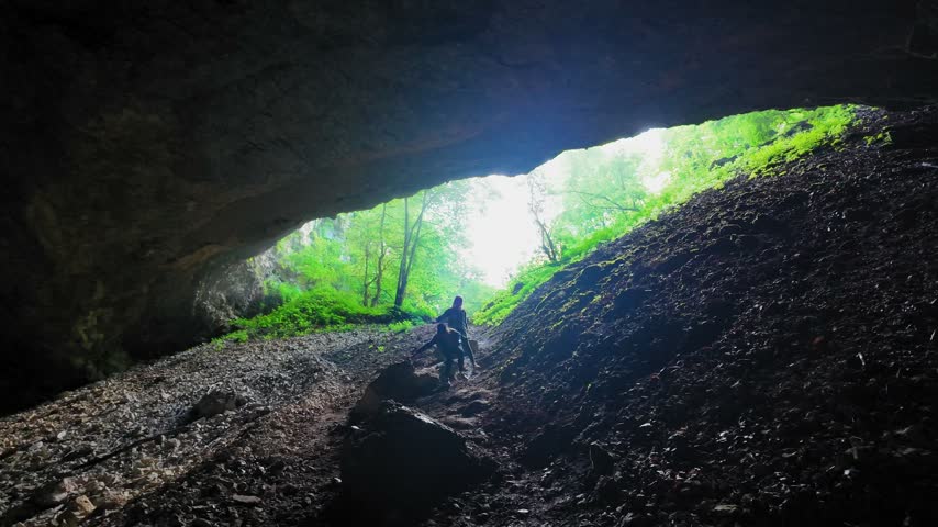 Two adventurous tourists explore the dark Sakinule Cave in Georgia, climbing a steep rocky path to a bright, sun-drenched entrance, discovering a beautiful green forest on a sunny day